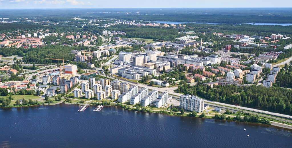 På denna bild ser du områdena Öbacka, Östra station, NUS (Norrlands universitetssjukhus) och Umeå älven. Älven slingrar sig genom staden och reflekterar det klara blåa himlen ovanför.